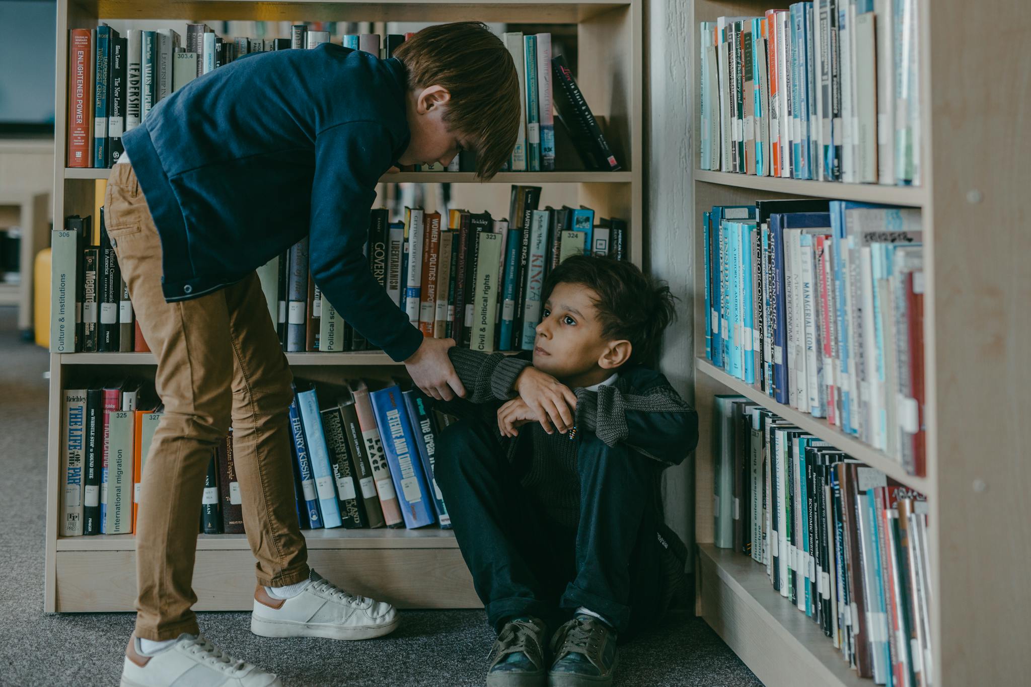 A concerned boy interacting with a classmate sitting on the floor in a library.