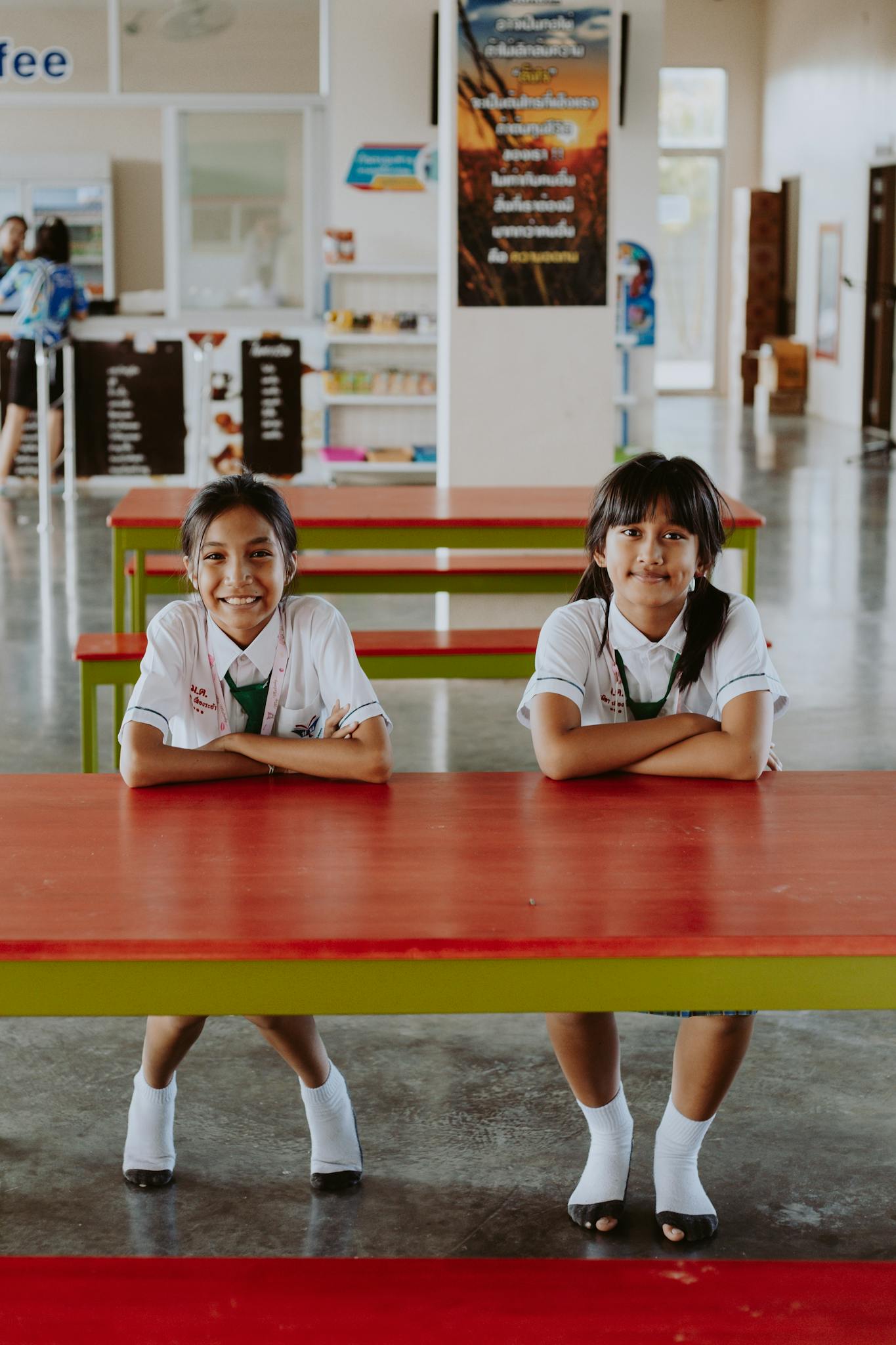 Two smiling Asian girls in school uniforms sitting at a red cafeteria table indoors.